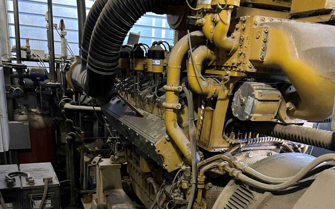Large yellow diesel engine with a curved black intake hose and numerous pipes in a dusty industrial room.