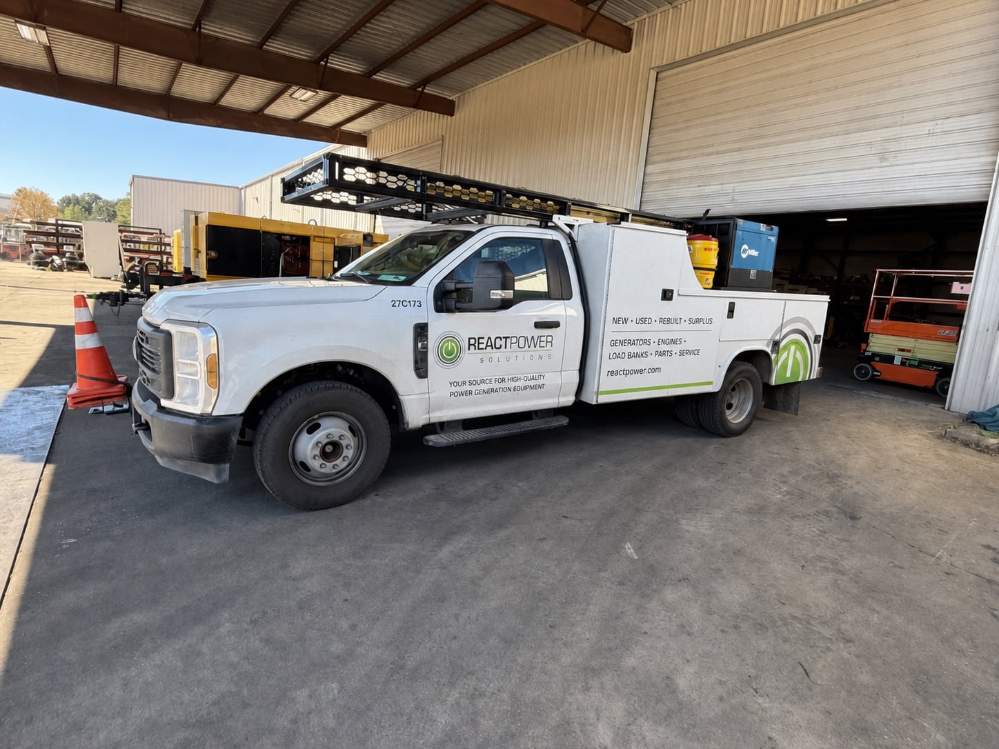 White service pickup truck with a ladder rack and tool/storage compartments parked in a warehouse bay, branding reads 'React Power Solutions'