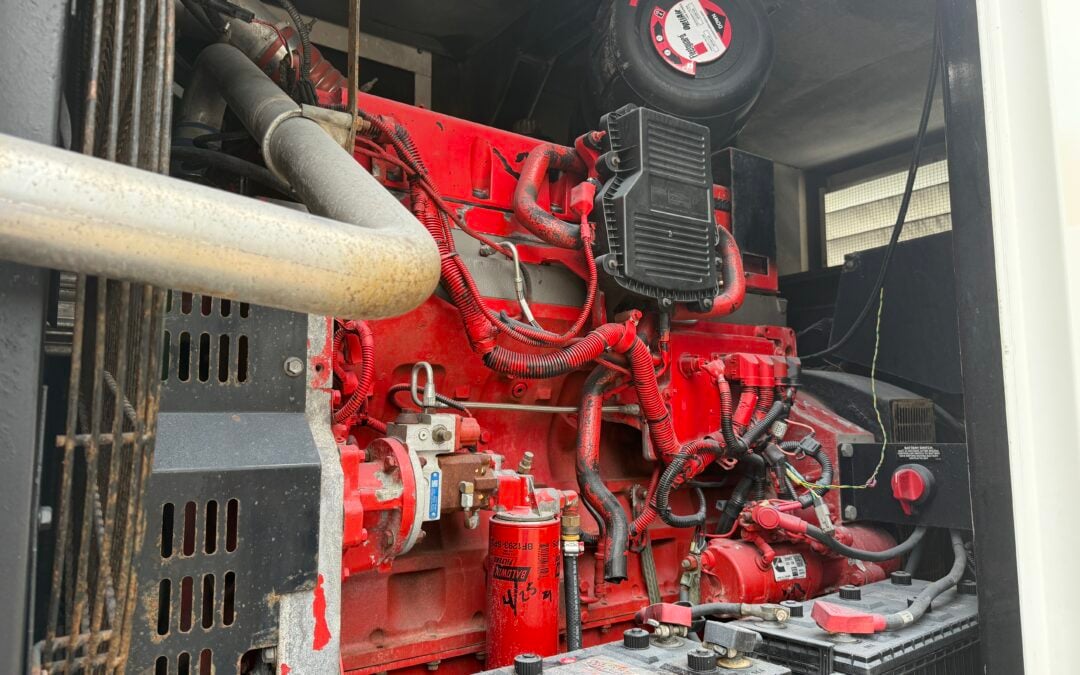 Red industrial engine with numerous hoses, wires, and a black control module in a machine cabinet. Battery bank visible at lower right.