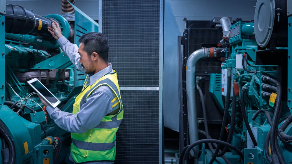 Maintenance technician in a high-visibility vest inspects a large industrial generator, using a tablet for monitoring.